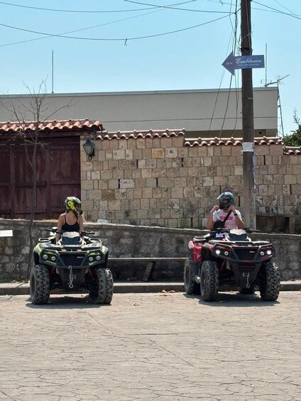 Two people on ATVs on a paved road with a stone wall and a building with a tiled roof in the background. 4