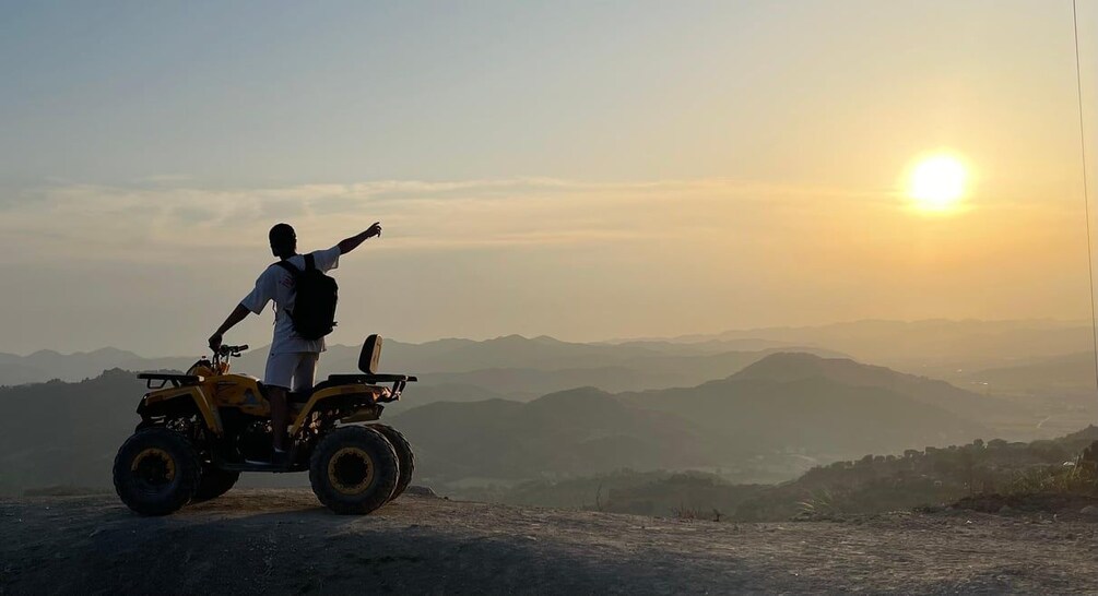 A person on a yellow ATV with a backpack, pointing towards the sun on a mountain. 2