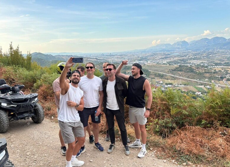 A group of people taking a selfie on a dirt path with a view of a city and mountains in the background. 1