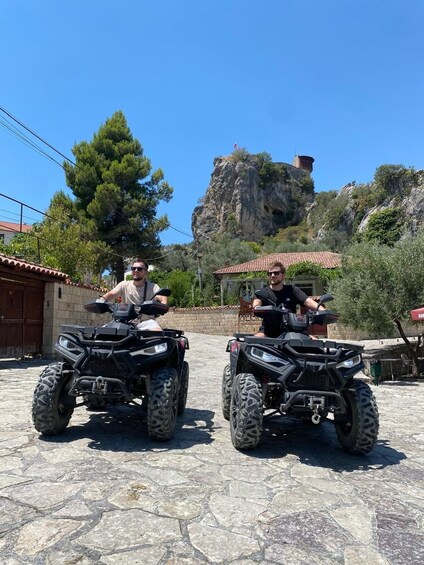 Two people on ATVs in a stone-paved area with a rocky hill and a red-roofed building in the background. 3