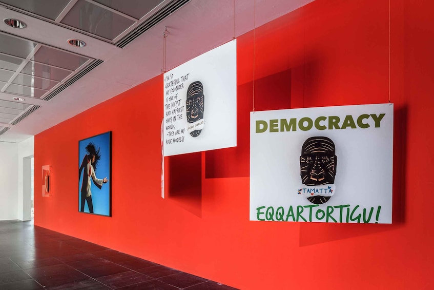 A red wall with three framed artworks, one of which has 'DEMOCRACY' and 'TAMATTA' text. 4