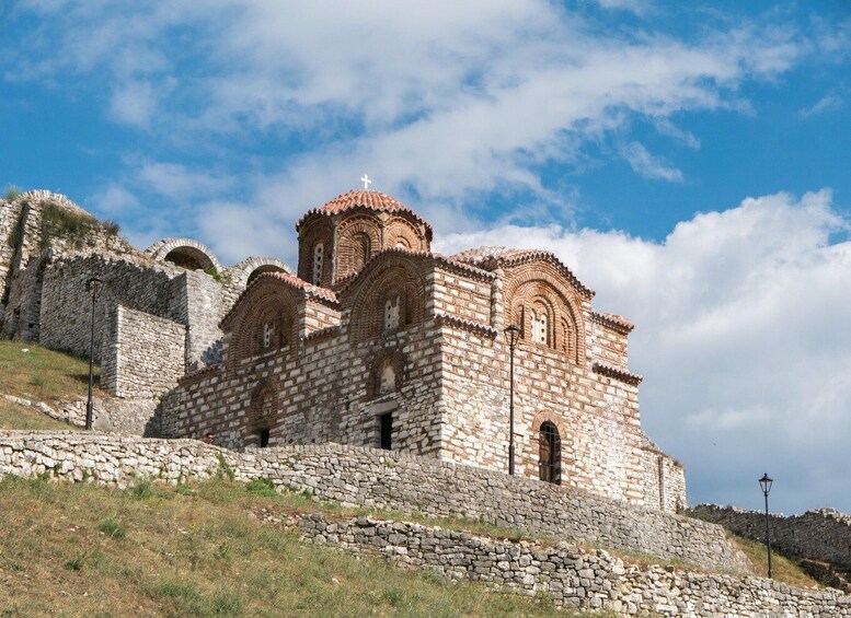 A stone church with multiple domes and a cross on top, surrounded by a stone wall and grassy hill. 5