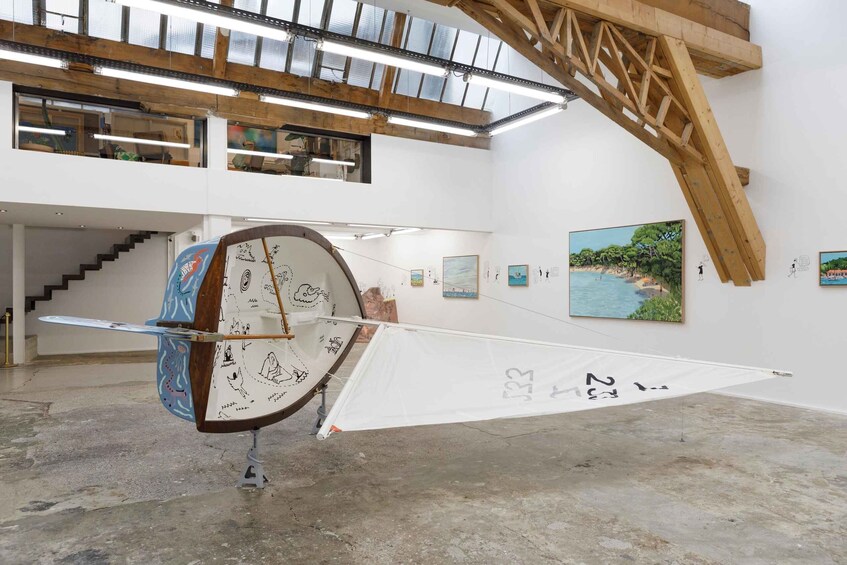 An art installation in a gallery with a large sail-like structure, paintings on the walls, and a staircase. 3