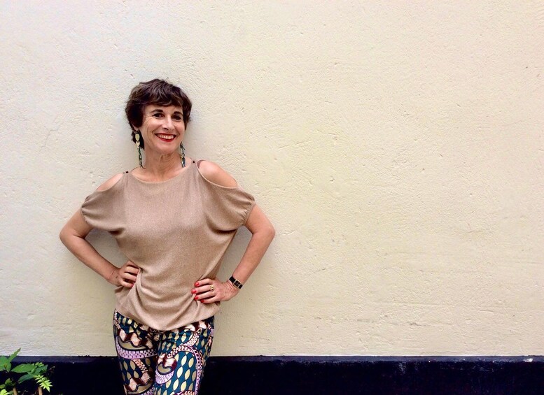 A person standing against a plain wall wearing a beige top and patterned pants. 4