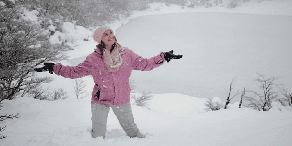 A person in a pink jacket and hat standing in a snowy landscape with trees and a body of water in the background. 3