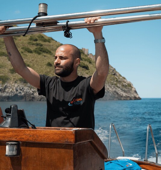 A man on a boat holding onto a metal railing, with a rocky coastline and calm sea in the background. 1