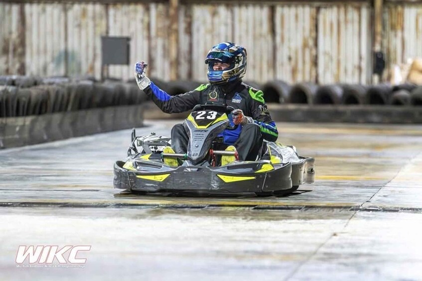 A go-kart racer in a black and yellow suit with the number 23, raising a hand, on a go-kart track. 1