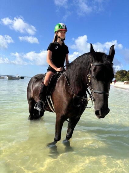 A person riding a horse in shallow water near a beach with boats in the background. 1