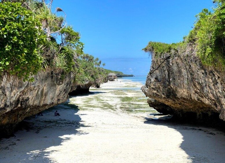 A beach with white sand, clear blue water, and rocky cliffs with greenery. 1