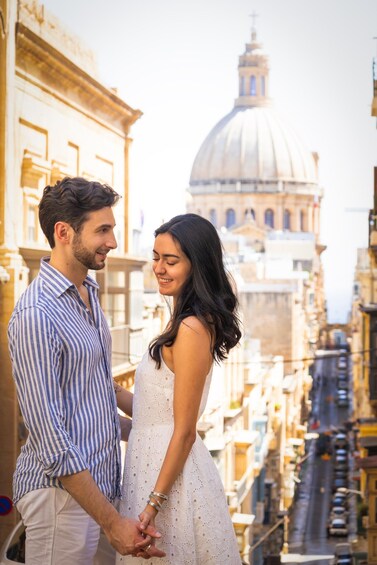 A couple holding hands on a balcony overlooking a cityscape with historic buildings and a prominent dome structure in the background. 3