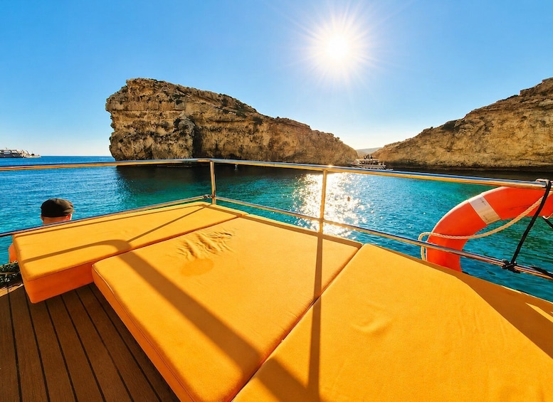 A sunlit deck with yellow cushions, a lifebuoy, and clear blue water with rocky cliffs. 4