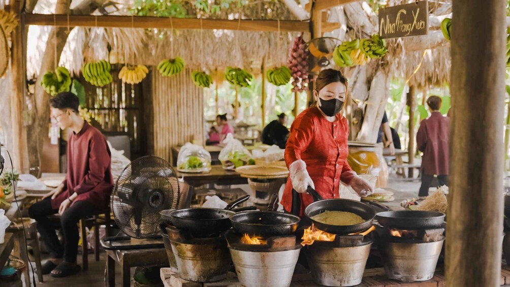 Un venditore di cibo di strada cucina all'aperto con pentole su fiamma viva, circondato da banane appese e in un ambiente rustico. 1