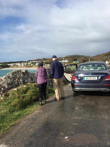 Two people standing by a parked car on a coastal road with houses in the background. 3