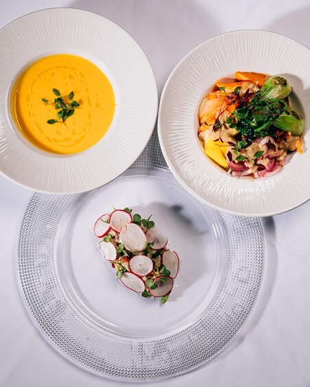 Three white plates with different dishes: a bowl of soup, a plate with vegetables and greens, and a plate with radishes and greens. 5