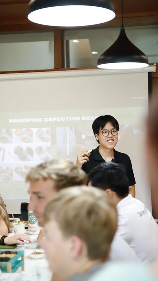 A presentation with a person holding a marker, a projector screen displaying 'HANDPICK DEFECTIVE BEAM', and a group of people seated at a table with food and drinks. 3