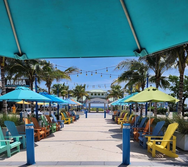 A beachside seating area with colorful Adirondack chairs and umbrellas, palm trees, and a sign reading "LAUDERDALE BY THE SEA". 1