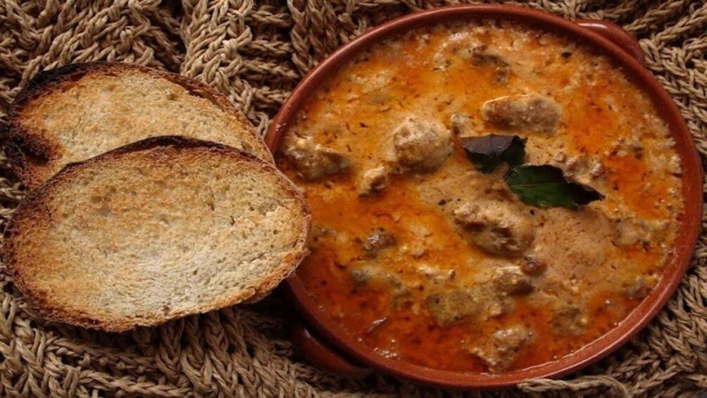 En skål suppe med frikadeller og pynt, ledsaget af ristede brødskiver på en vævet måtte. 1
