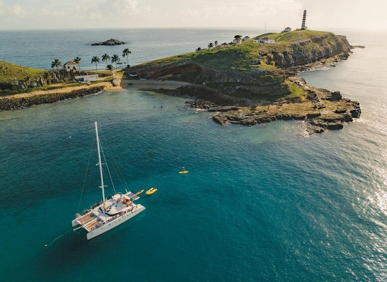 A sailboat anchored near a rocky coastline with a lighthouse and palm trees. 1