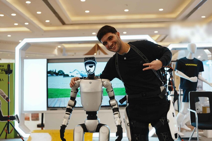 A person standing next to a humanoid robot in a modern indoor setting with a large screen displaying a landscape in the background. 1