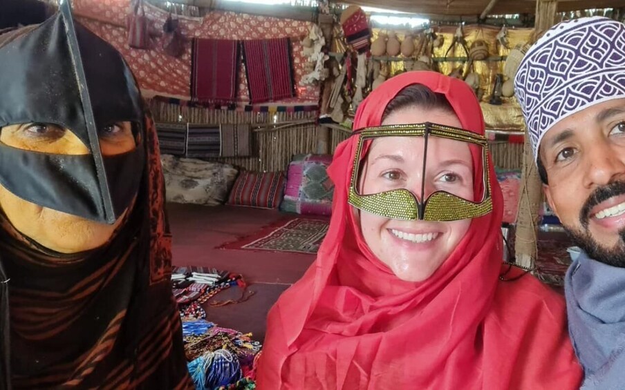 A person in a red headscarf and another in a patterned headscarf, with a background of textiles and rugs. 5