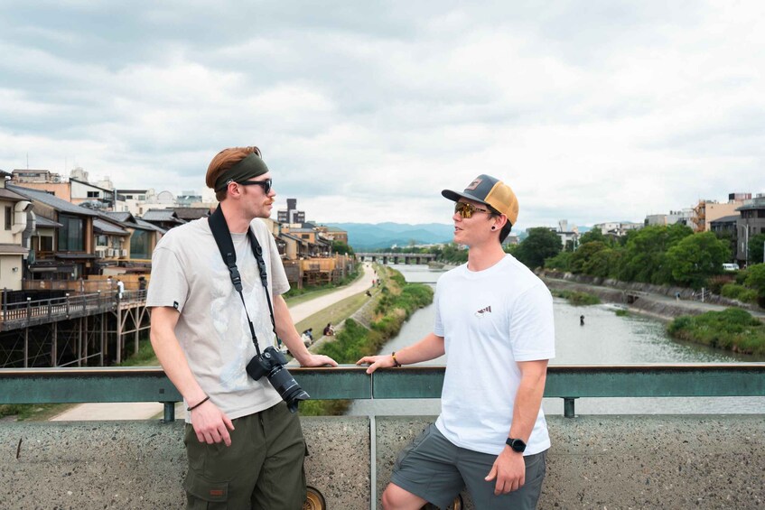 Two people standing on a bridge overlooking a river with buildings and greenery in the background. 3