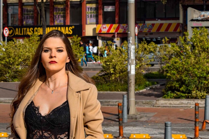 A woman in a tan coat and black lace top standing in an urban street with shops and greenery in the background. 2