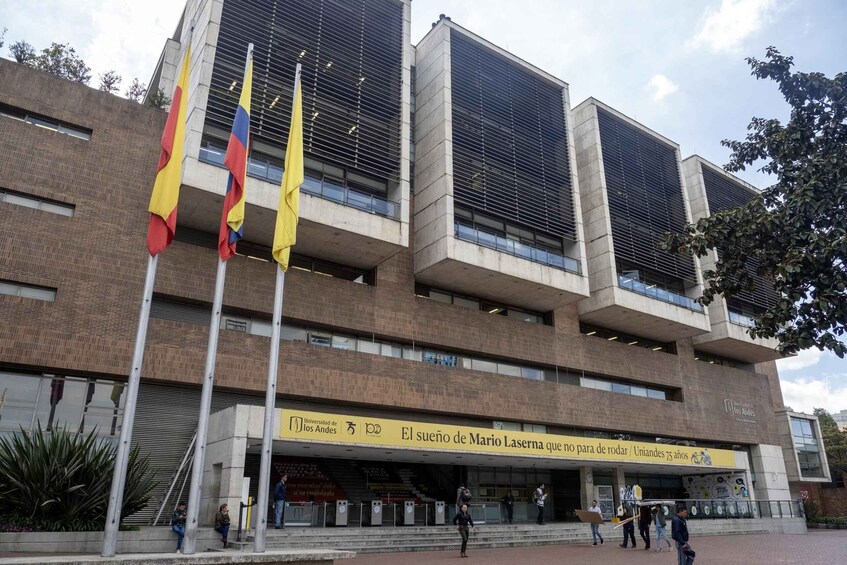 A modern building with multiple floors and large windows. There are several flags in front of the building, and a banner with text is displayed on the lower part of the facade. 3