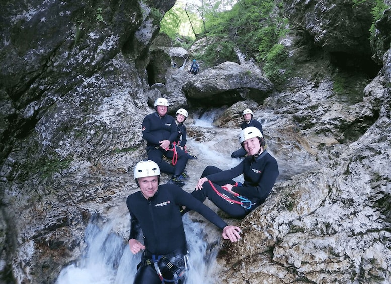 A group of people in wetsuits and helmets sitting on a rocky ledge by a flowing waterfall. 5