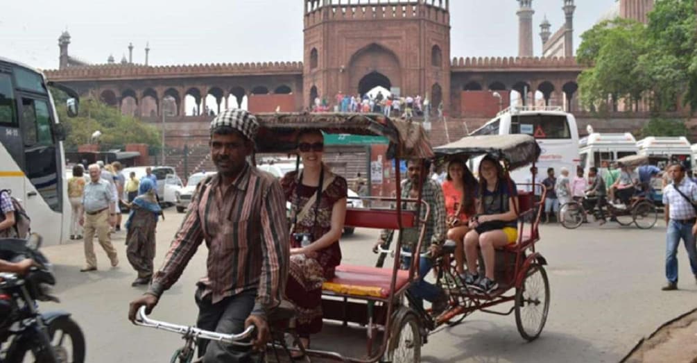 A man pushing a rickshaw with passengers, a large red building with arches in the background, and a crowd of people. 5