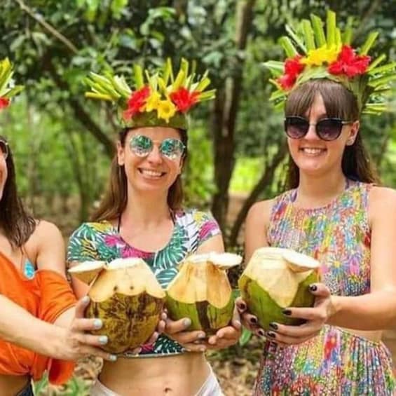 Three people holding coconuts with decorative headpieces in a forested area. 1