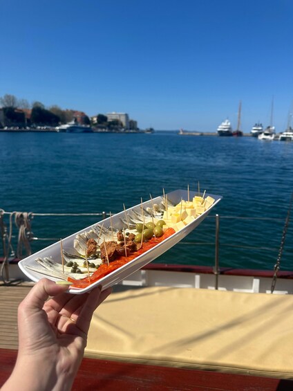 A hand holding a plate of food with various items including olives, cheese, and meats, against a backdrop of a body of water with boats and buildings. 4