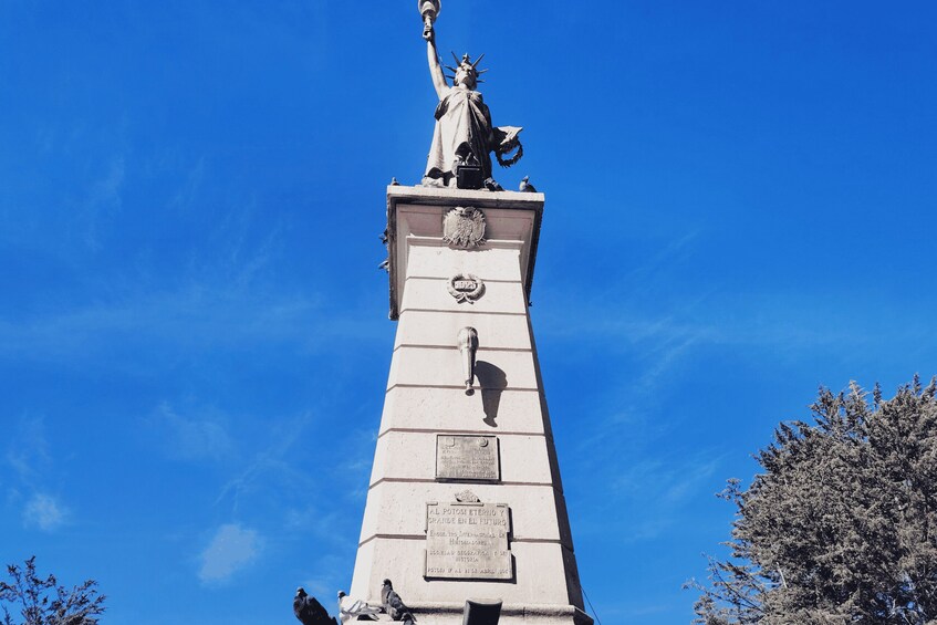 A tall monument with a statue of a robed figure holding a torch, surrounded by birds. 2