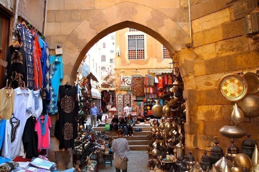 A bustling market with a stone archway, various stalls selling textiles and metal items, and a busy street in the background. 5