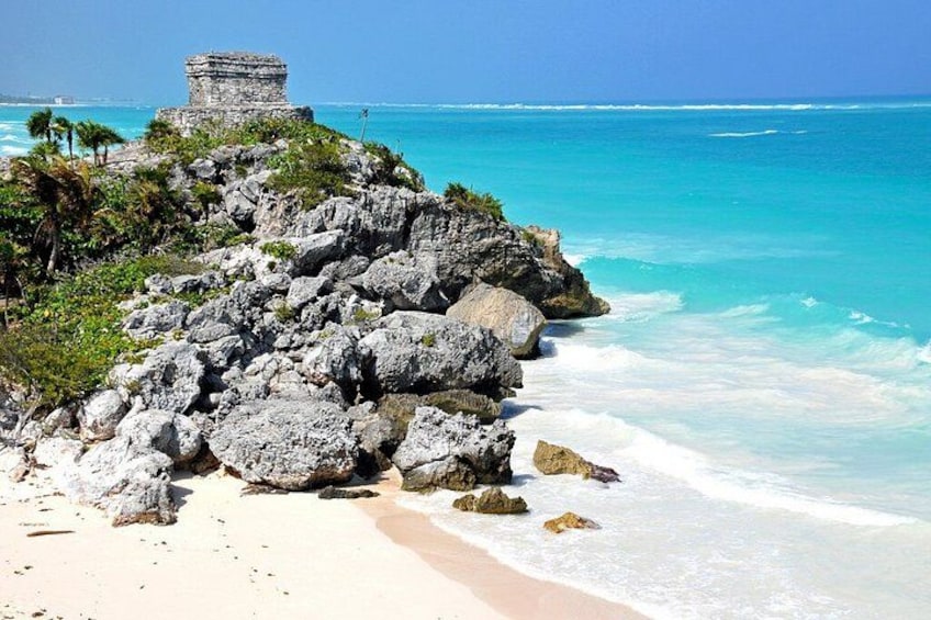 A coastal landscape with a rocky outcrop, a small ancient structure, and turquoise ocean waves. 1