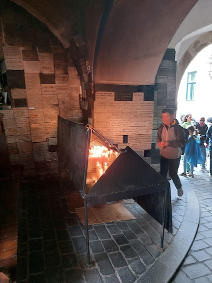 A stone wall with inscriptions, a metal structure with candles, and a person walking by. 4