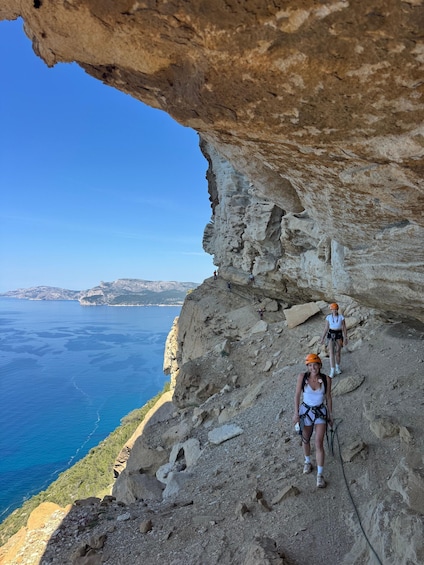 Two people climbing a rocky cliff with a clear view of the sea and distant hills. 2