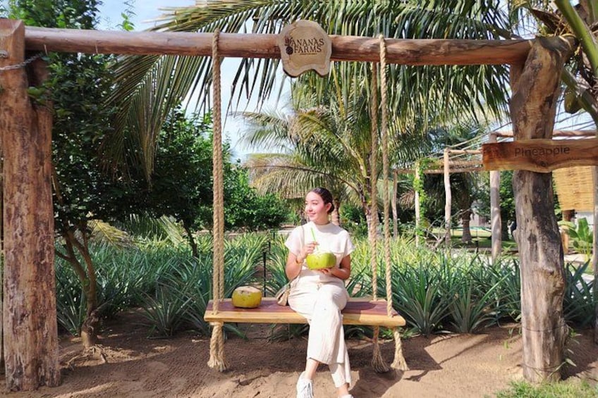 A woman sitting on a wooden swing in a tropical garden with a sign reading 'Nanas Farms.' 1