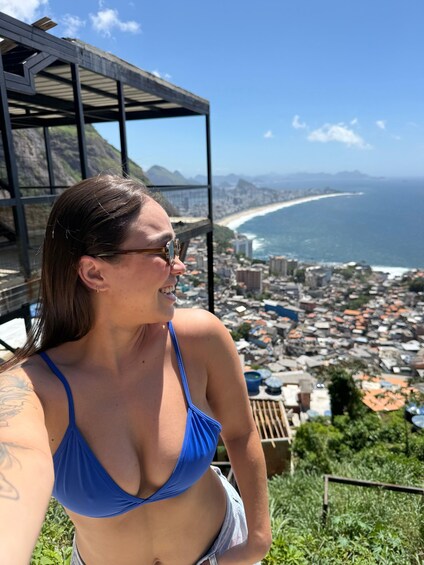A person in a blue bikini top and white underwear, with a tattoo on their arm, overlooking a coastal city with a beach and mountains in the background. 5