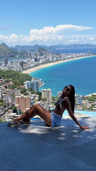 A person sitting on a ledge overlooking a coastal city with a beach and mountains in the background. 2
