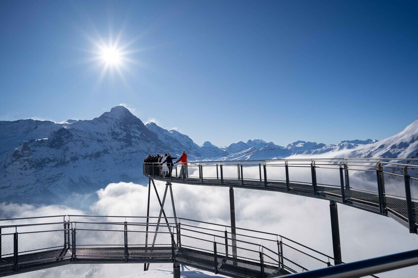 A metal walkway with people walking above clouds, overlooking snow-covered mountains under a clear sky. 4