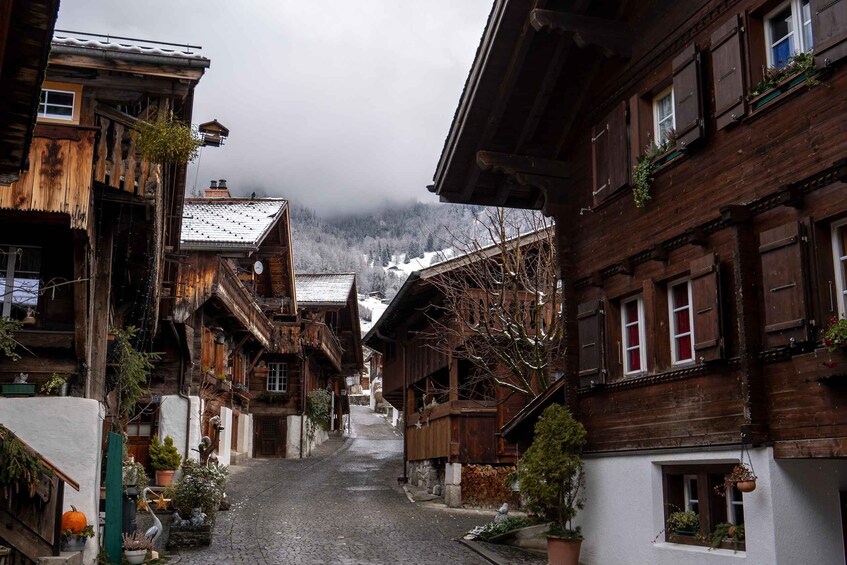 A narrow cobblestone street lined with wooden houses, some with balconies and flower boxes, leading towards a snowy mountain. 1