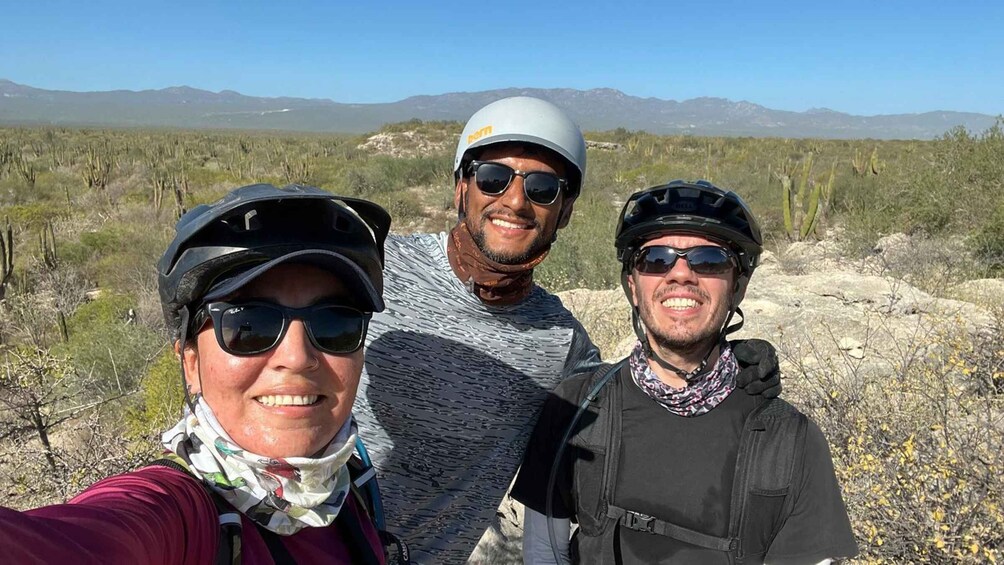 Three people wearing helmets and sunglasses, standing in a desert landscape with sparse vegetation and mountains in the background. 5