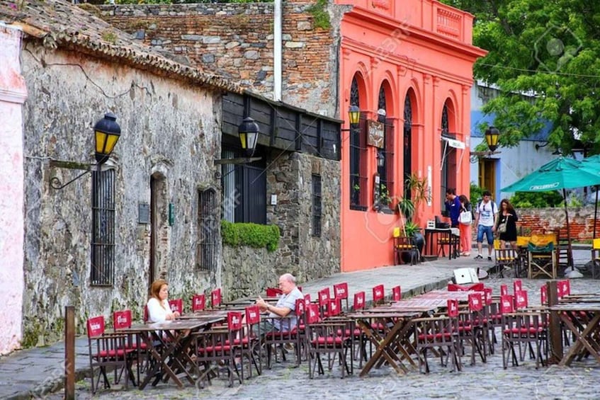 Outdoor tables and chairs in a cobblestone street, a stone building, and a red building with arches. 3