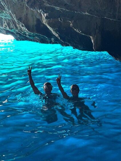 Two people swimming in clear blue water with a rocky cave in the background. 1
