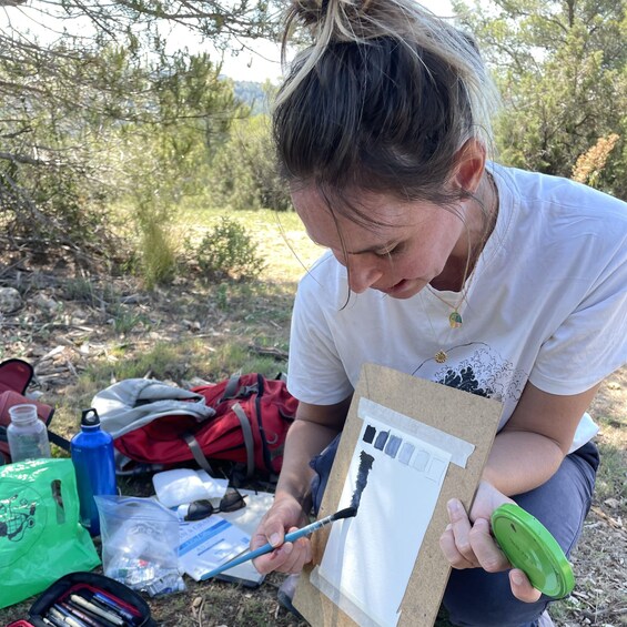 A person painting on a canvas outdoors with a backpack and art supplies nearby. 2