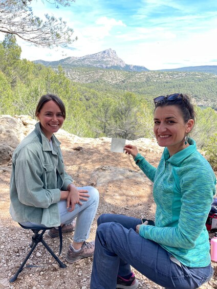 Two people sitting on a tripod stool outdoors with a mountainous landscape in the background. 3