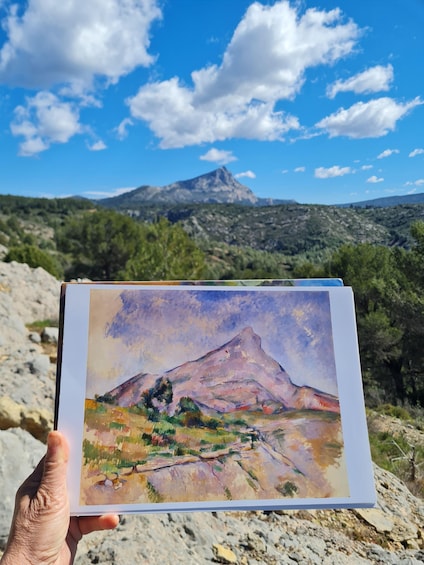 A hand holding a painting of a mountain landscape with a clear sky. 2