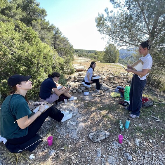 A group of people sitting and standing outdoors, some holding notebooks and pens, in a natural setting with trees and rocks. 5