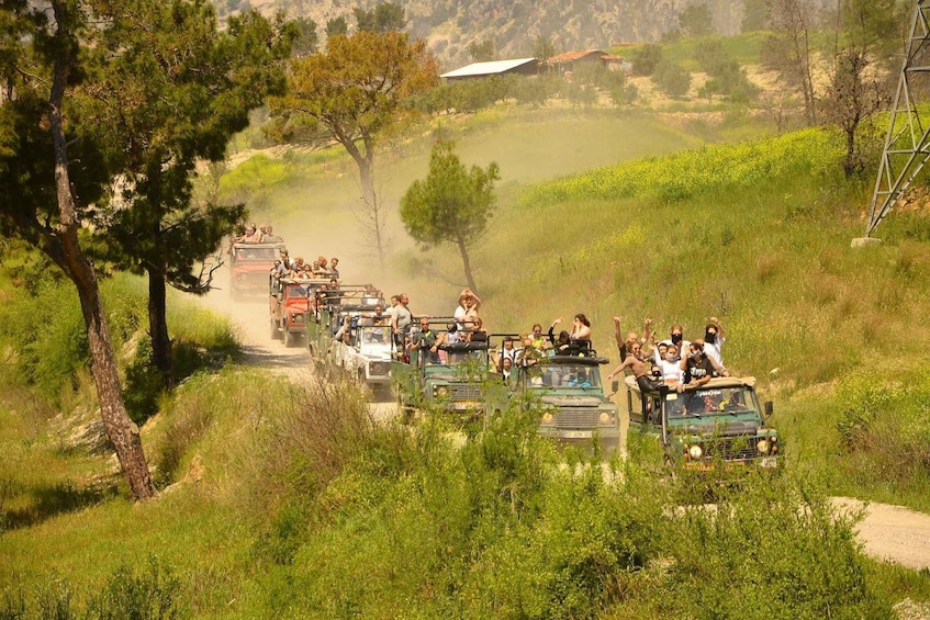 A convoy of off-road vehicles with passengers on a dirt road surrounded by greenery and hills. 1