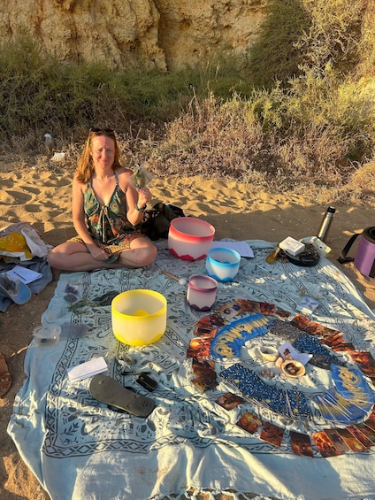 A person sitting on a blanket with tarot cards, bowls, and a thermos on a sandy beach. 2